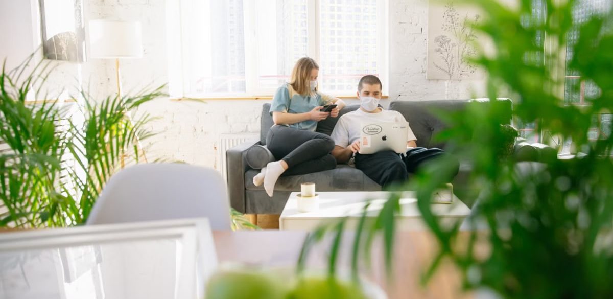 Couple sitting on sofa with masks, using electronics in bright room.