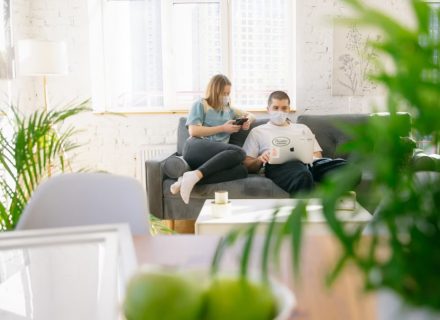 Couple sitting on sofa with masks, using electronics in bright room.
