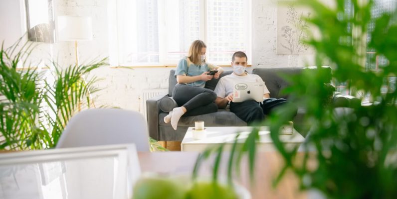 Couple sitting on sofa with masks, using electronics in bright room.